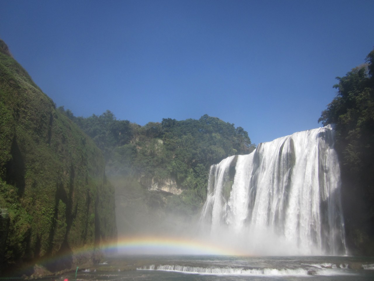 Huangguoshu Main Waterfall pours over the cliff, with a rainbow in the spray and a blue sky.