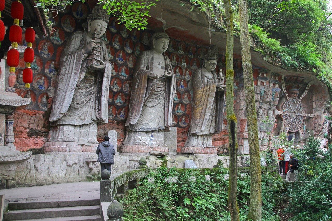 Large standing Buddhist figures carved into a painted cliff at Baodingshan, Dazu, with a visitor below showing their scale.