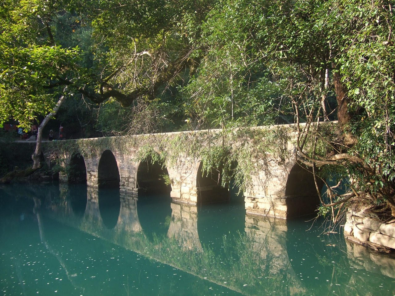 The old stone-arch Xiaoqikong bridge reflected in blue-green water, surrounded by dense trees in Libo.