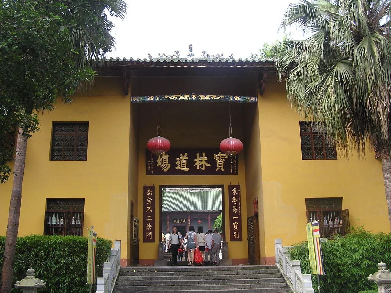 The yellow gate building of Nanhua Temple in Shaoguan, with red lanterns, a dark plaque, trees and visitors on the steps.