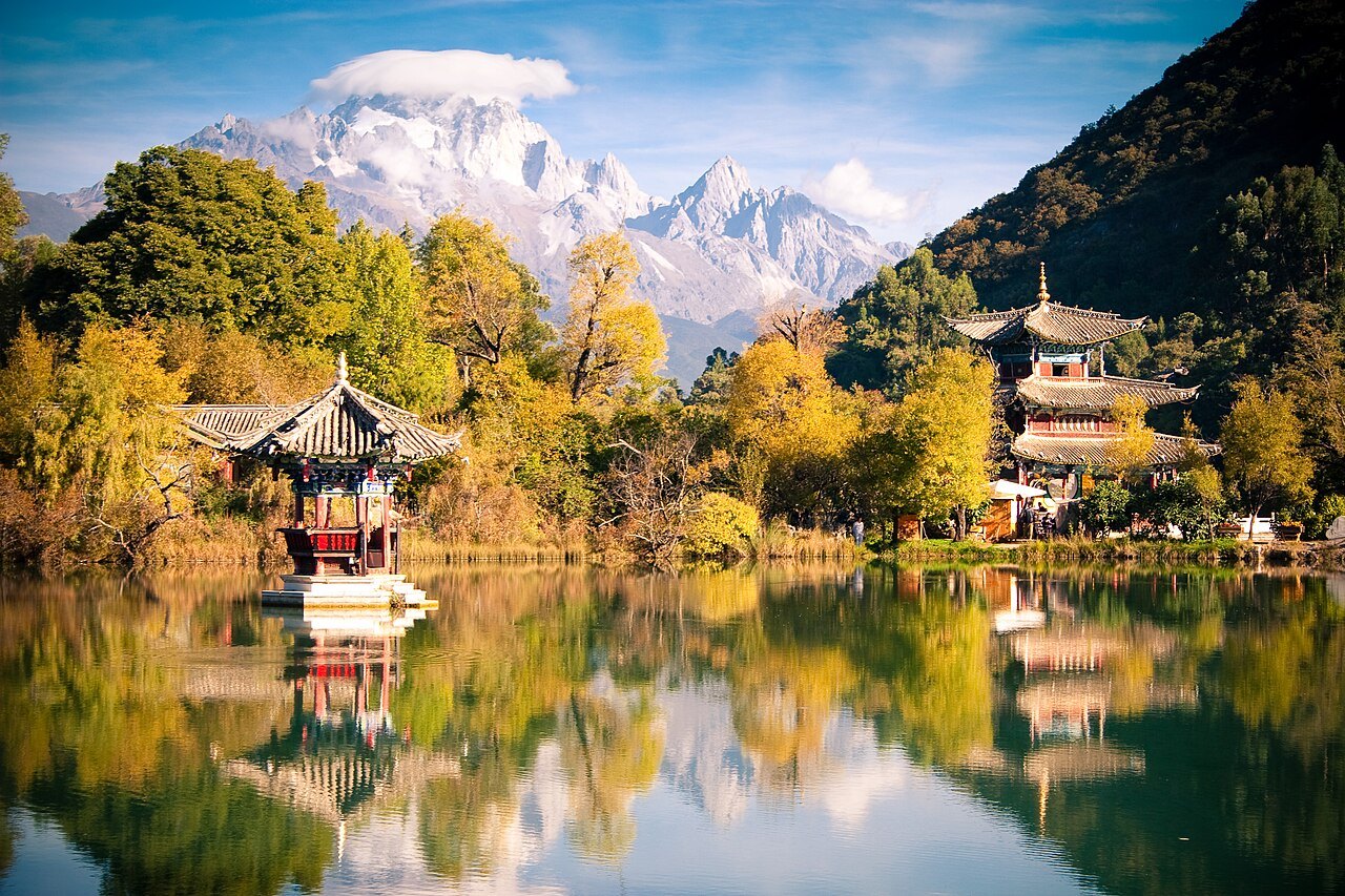 Pavilions and autumn trees reflected in Black Dragon Pool beneath snow-covered mountains.