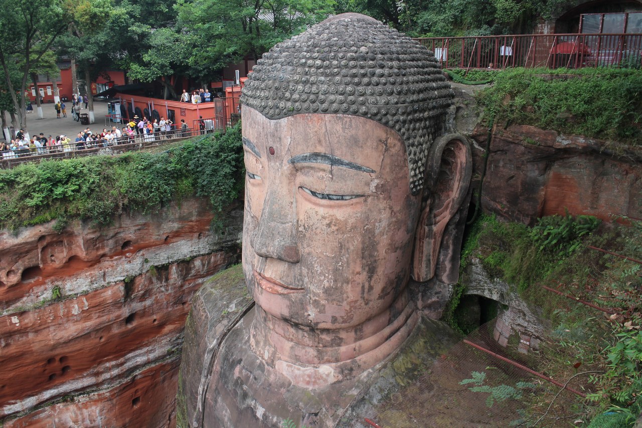 Close view of the Leshan Giant Buddha’s head, with red rock, trees and visitors showing its scale.