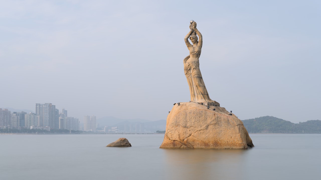 The Fisher Girl statue rises from a granite boulder in Xianglu Bay, with Zhuhai buildings behind it.