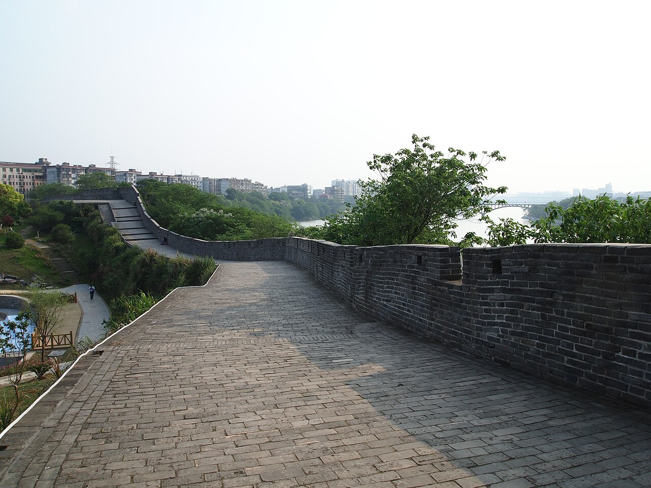 A brick city-wall walkway beside trees and the river near Yugu Pavilion in Ganzhou.
