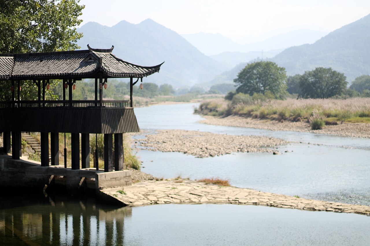 A riverside wooden pavilion and wooded hills at Guyan Huaxiang, Lishui
