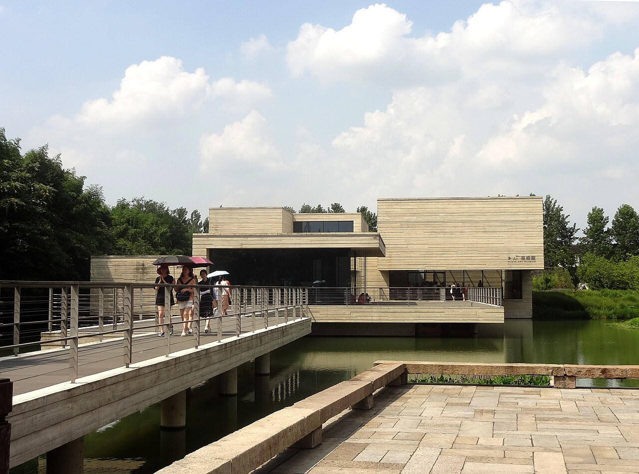 A footbridge approaches the low modern stone building of Mu Xin Art Museum over water